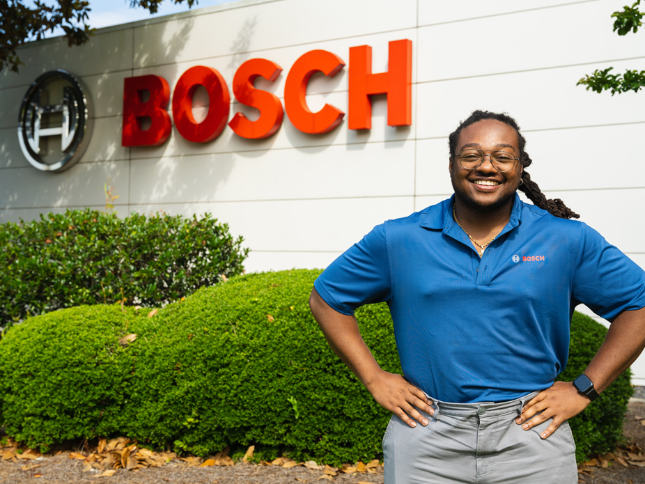 A smiling CofC student wearing a blue Bosch polo stands confidently in front of the Bosch company logo and neatly trimmed shrubbery.