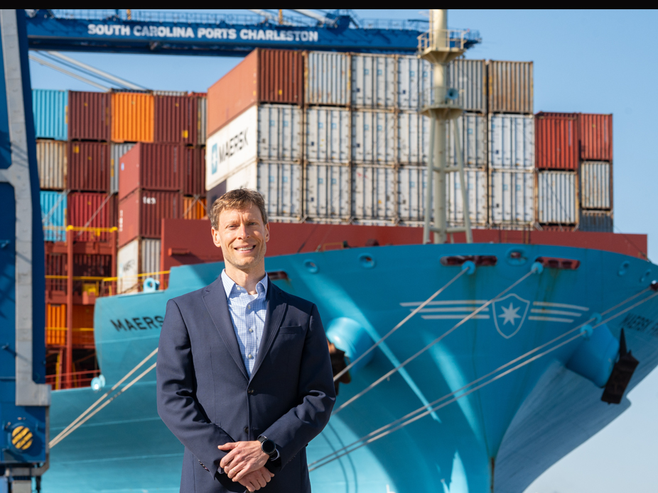 Micah Mallace smiles in a blue suit jacket and blue and white checkered shirt, stands at the South Carolina Ports Charleston with a ship loaded with containers in the background.