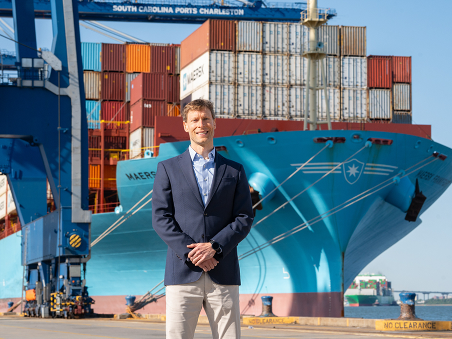 Alum Micah Mallace standing on the dock of the Port of Charleston with a large ship loaded with shipping containers in the background.