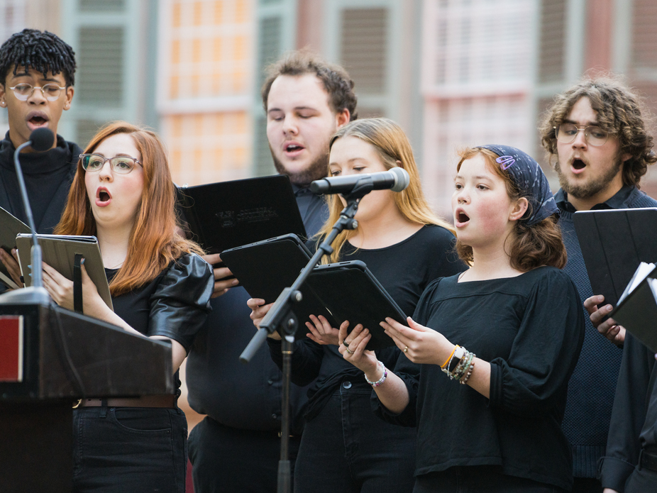 Group of singers performing outdoors, holding black music folders and singing into microphones.
