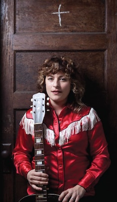 Cary Ann Hearst in a red fringed shirt holding a guitar, seated against a dark wooden door.