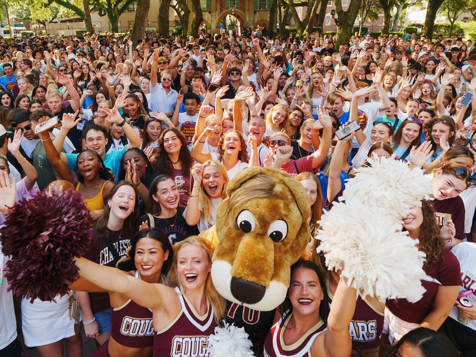 Large crowd of cheering college students gathered outdoors around Clyde the Cougar mascot and CofC cheerleaders holding pom-poms.
