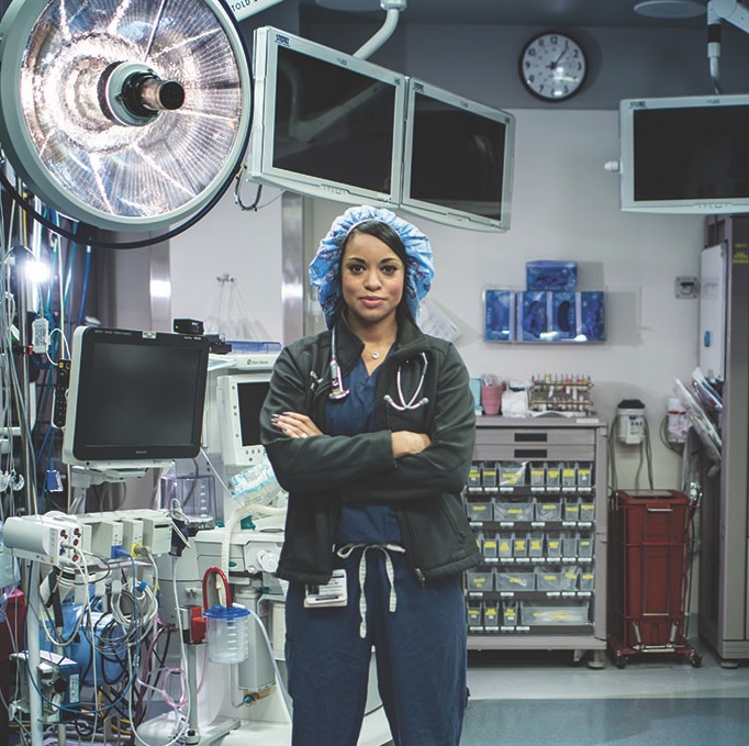 Dr. Hilton in scrubs with a stethoscope stands with arms crossed in an operating room surrounded by medical equipment.