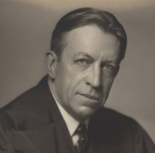 Black-and-white portrait of a J Waring in a suit, looking toward the camera.