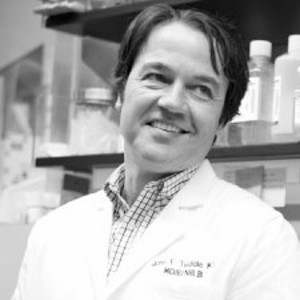 Dr. John Tisdale smiling in a lab coat standing in a laboratory with shelves of bottles in the background.