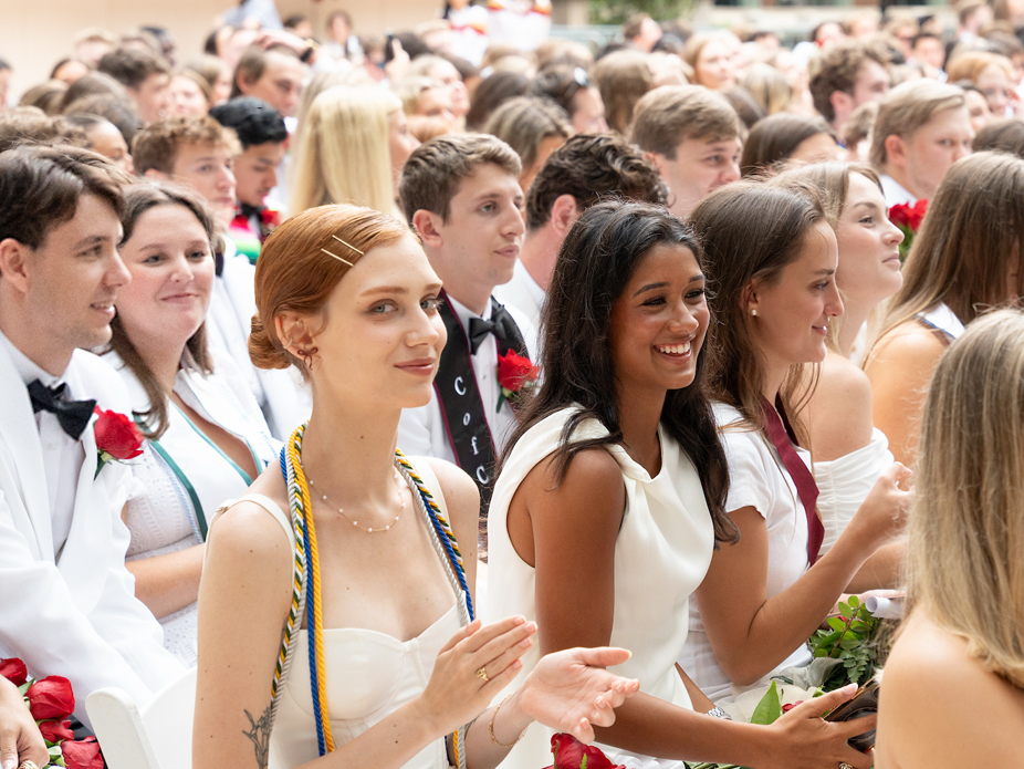 Seated group of College of Charleston graduating students in white outfits, smiling and applauding during an outdoor ceremony.