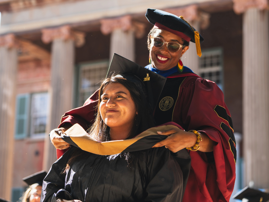 Graduate in cap and gown receives a ceremonial hood during commencement outside a columned campus building