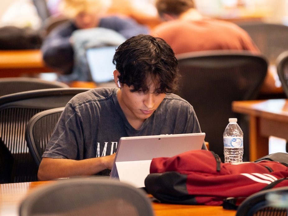 Student uses a tablet at a study table with a backpack and water bottle in a large classroom or library.