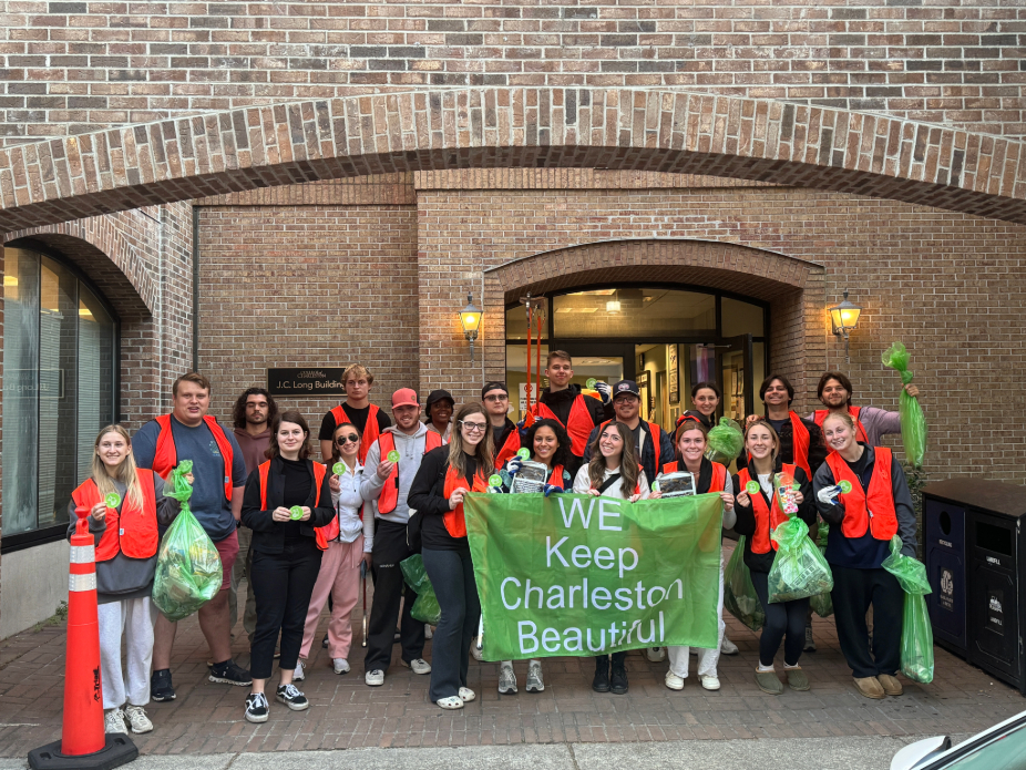 A group of students with orange vests and trash bags holding up a flag that says "We Keep Charleston Beautiful"
