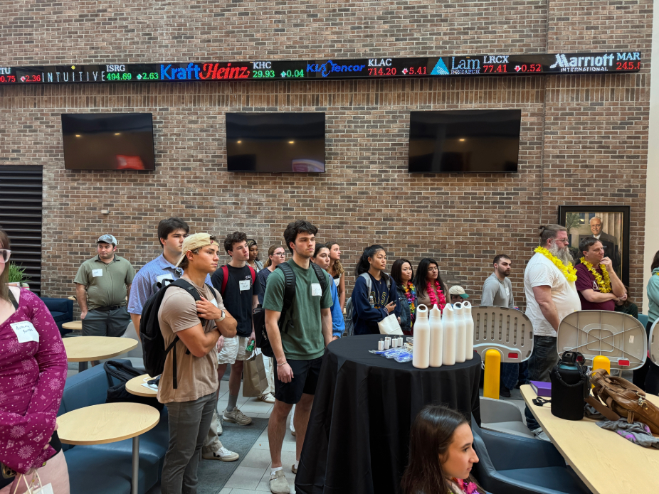 A group of people gathered in an indoor space with exposed brick walls, attending an event. Several round tables and chairs are arranged throughout the room. A black table with white bottles is positioned in the foreground. Above the group, an electronic ticker displays stock prices for companies like Intuitive, Kraft Heinz, KLA Corporation, Lam Research, and Marriott International. Three large screens are mounted above the ticker. Some attendees are wearing name tags.