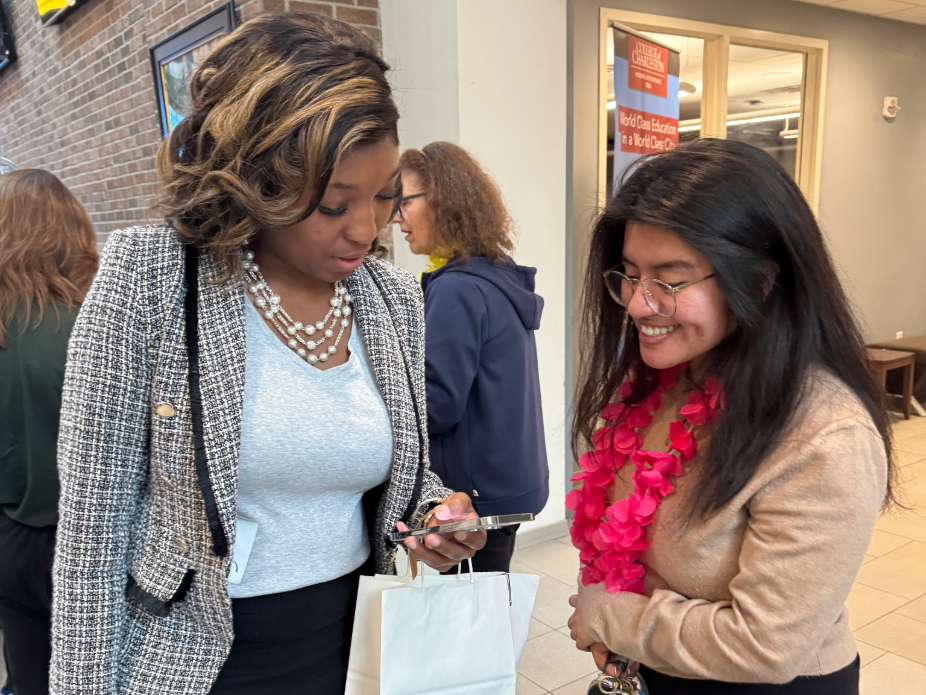 Two individuals conversing indoors; one holds a white shopping bag and looks at a smartphone, while the other wears a pink lei.