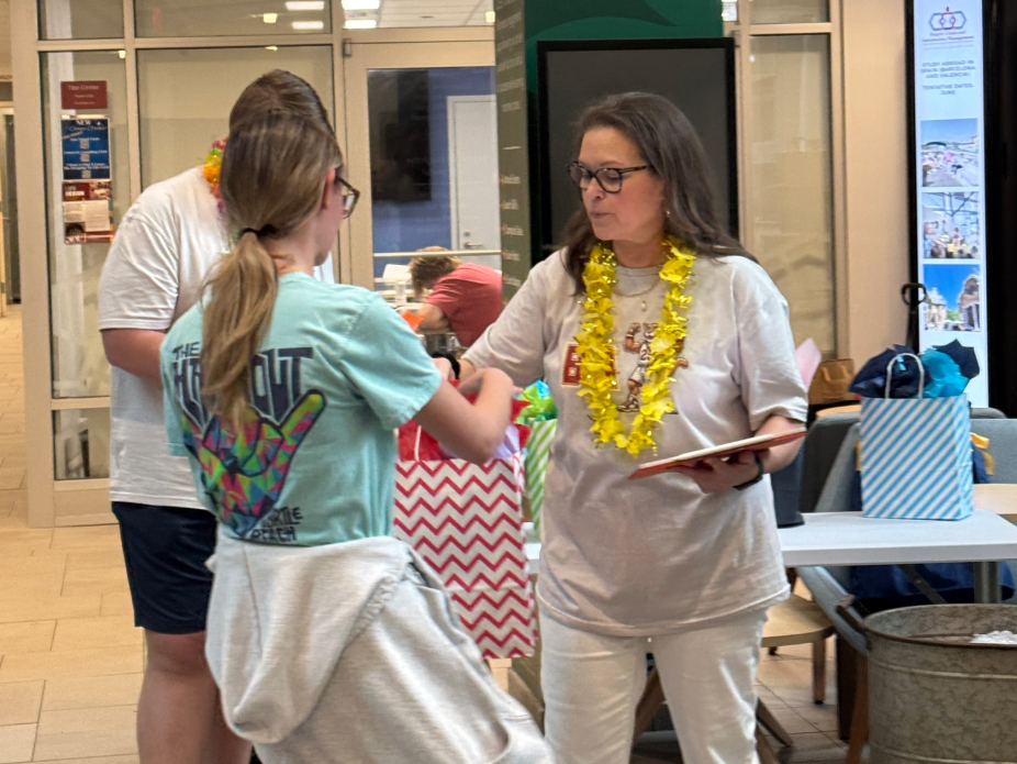 Three people stand in a lobby or common area during a celebratory event. Two are facing each other—one holding a gift bag, the other a folder or book—and both wear leis. A third person stands nearby in casual attire. Tables with gift bags and decorations are visible in the background. 