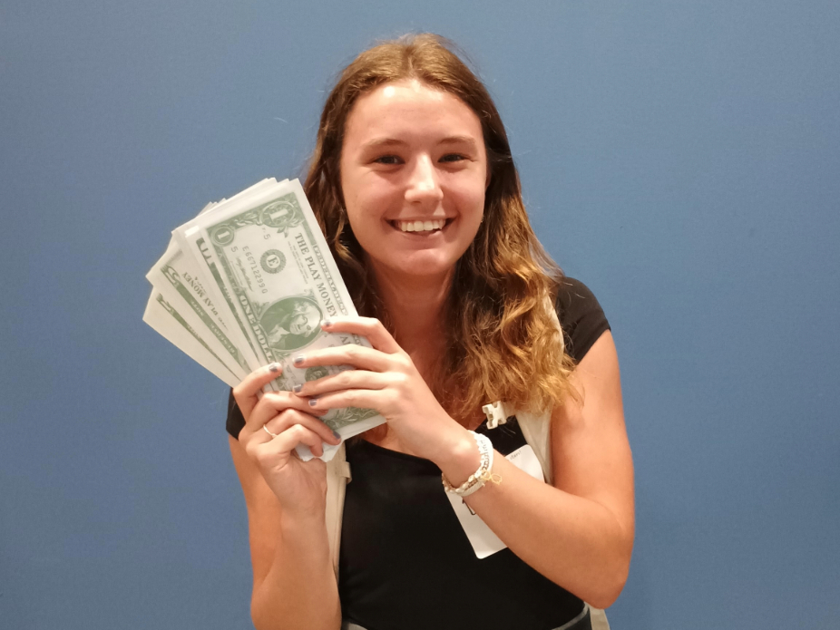 A person with long hair, dressed in a black shirt and white apron, holds several one-dollar bills in both hands. The background is a plain blue wall.