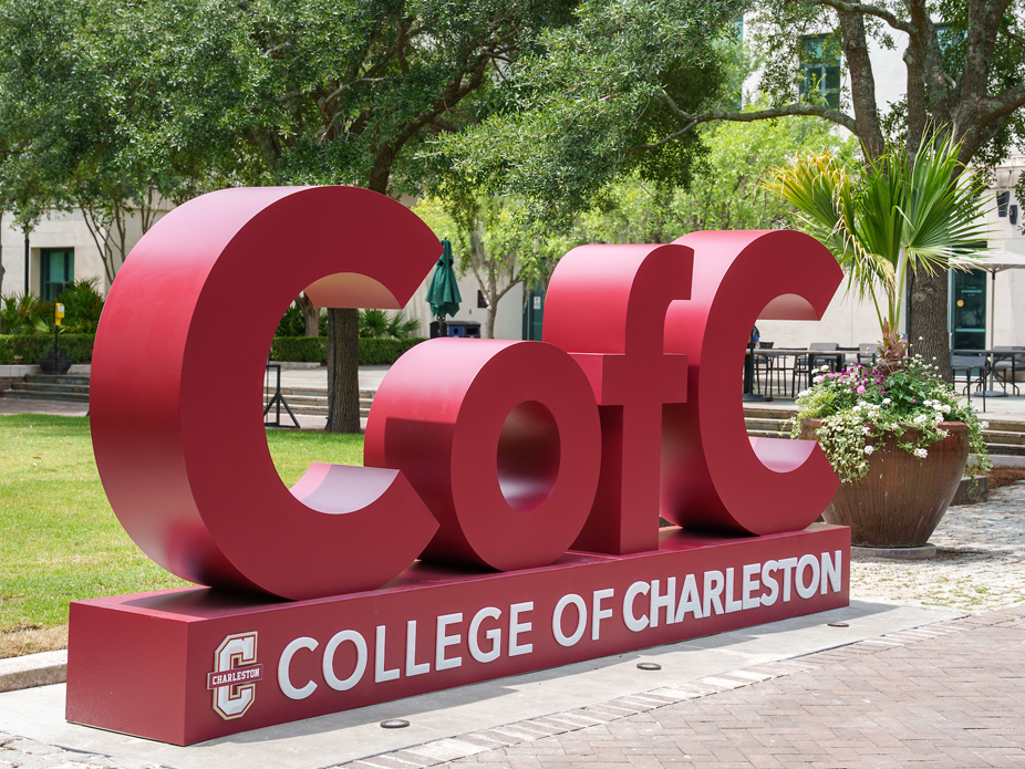 Large red “CofC” sculpture on a campus lawn with a base reading “College of Charleston,” surrounded by trees and outdoor seating.