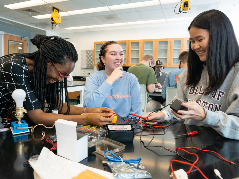 Students collaborate at a lab bench, smiling while testing an electrical circuit with wires and a multimeter in a classroom lab.