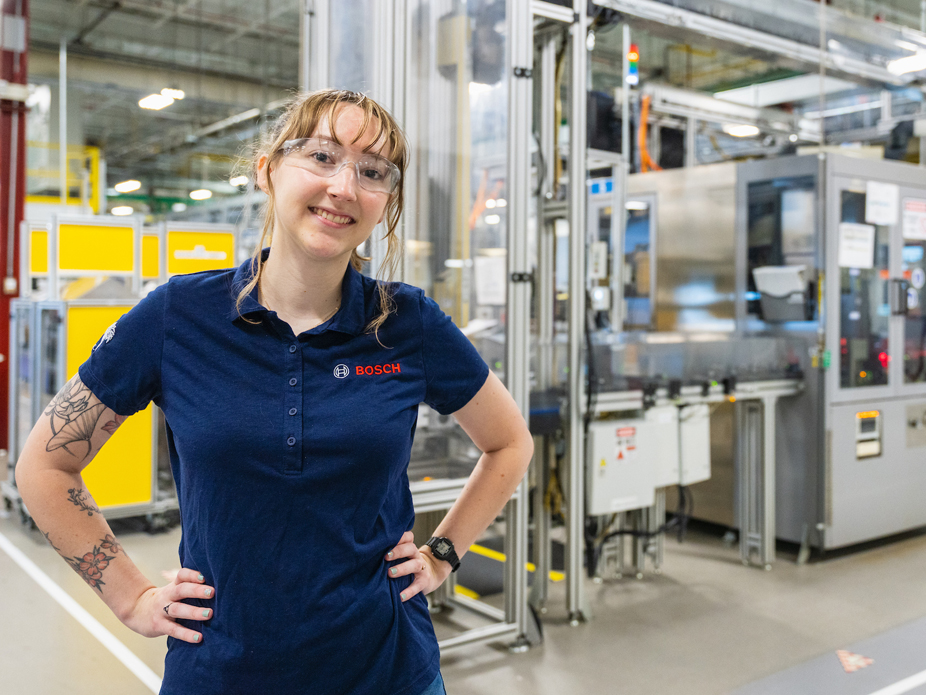 Portrait of Ashley Dowd, a smiling young woman with hands on her hips wearing safety glasses and a "Bosch" polo shirt with engineering equipment in the background.