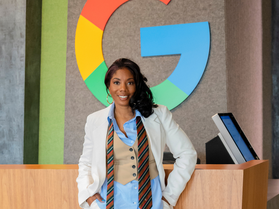 Profession portrait of Rachel Olden smiling wearing a suit with un-ied tie around her neck in front of the Google "G" logo.