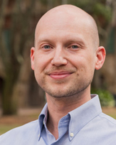 A professional headshot of a Robert Bux with a shaved head, wearing a light blue button-up shirt.