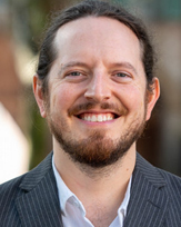 Aaron Hancock, a smiling man with a beard, wearing a striped suit jacket over a white shirt, in an outdoor setting.