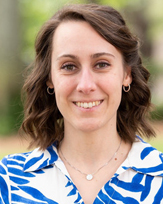 Professional headshot of Ana Lavado with curly hair, wearing a blue and white patterned shirt, smiling outdoors.
