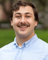 Tradd Tobin with a mustache smiles outdoors, wearing a light blue button-up shirt.