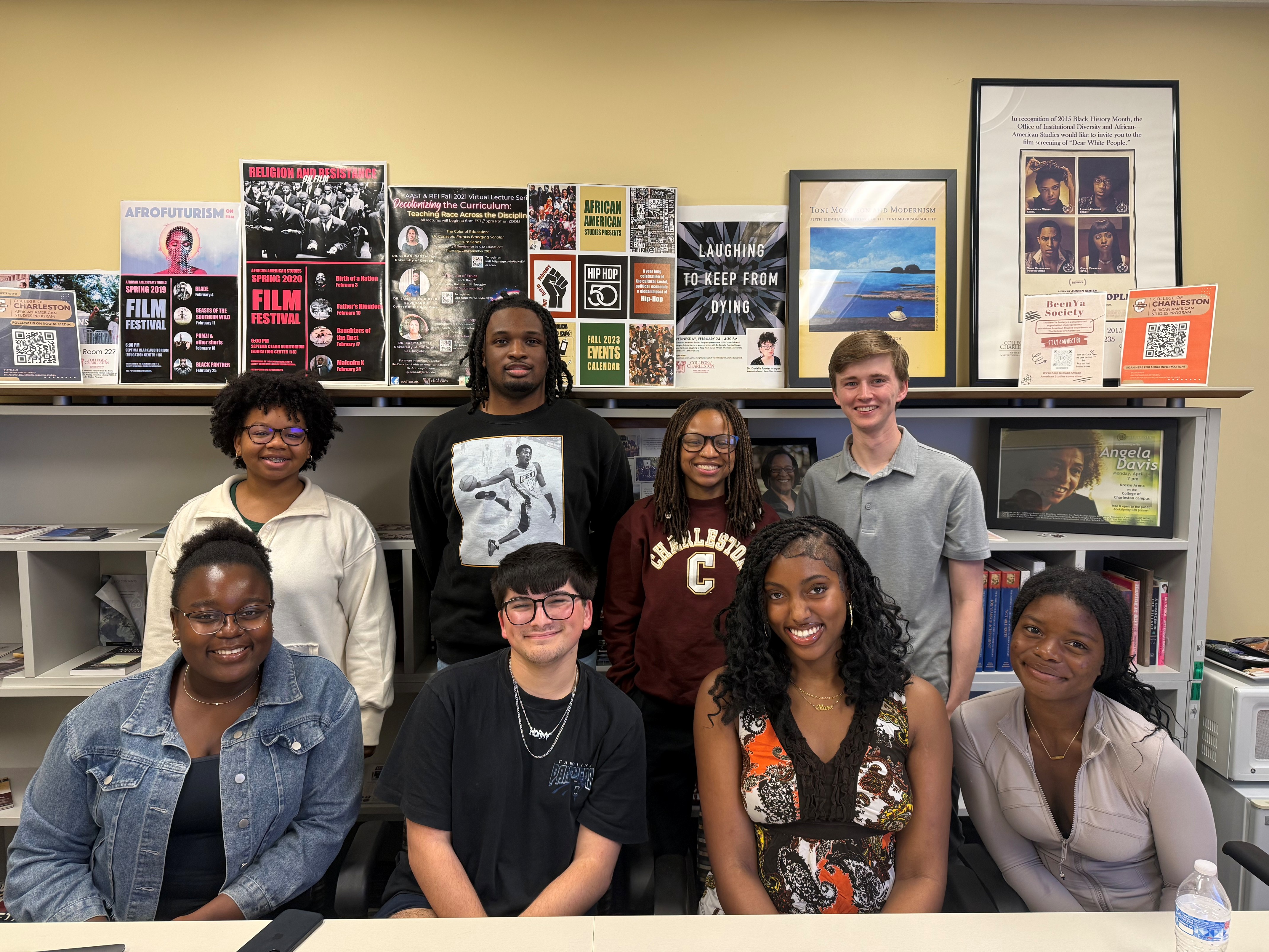 Group photo of ten people arranged in two rows in front of shelves and a wall covered with posters and framed prints. The background includes readable posters and flyers with text such as “Afrofuturism,” “Spring 2020 Film Festival,” “Hip Hop at 50,” and a framed poster featuring Angela Davis, creating an academic or cultural office setting.