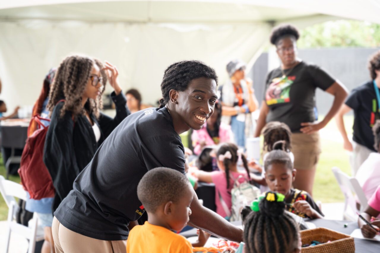  Person assists children with a hands-on activity at a community event under a large outdoor tent, with participants gathered around tables in the background.