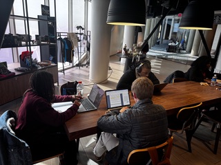 Three people work on laptops around a large wooden table in a bright, modern workspace with tall windows, hanging lights, and clothing racks in the background.