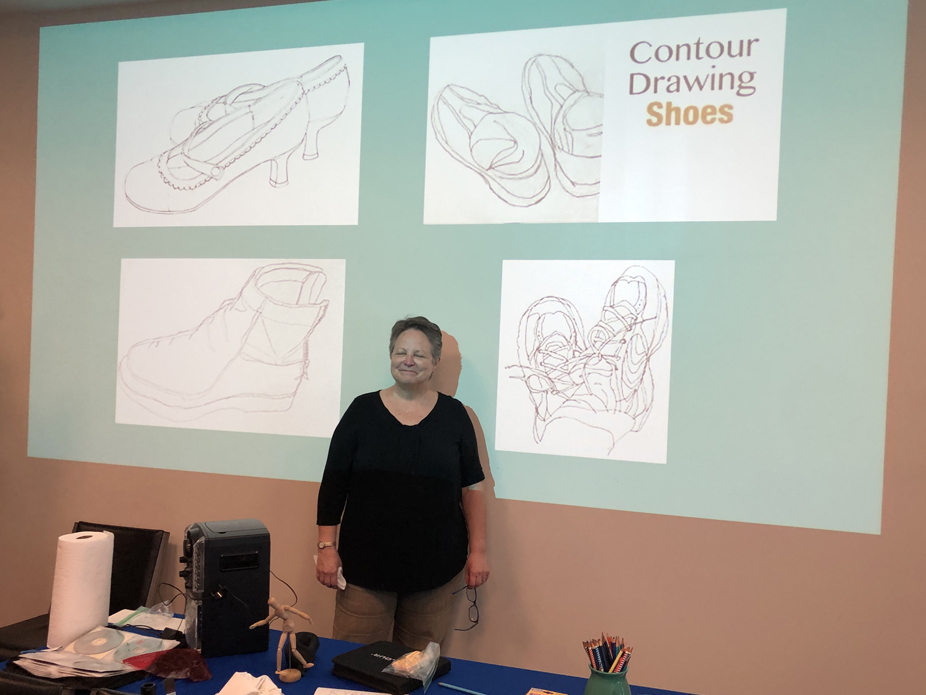 Artist Becky Sharpe stands before a screen showing drawings or shoes with the title of her topic "Contour Drawing Shoes"  of shoes 