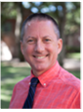 Professional portrait of a smiling man wearing a coral shirt and tie, set against a natural outdoor background.
