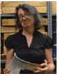 A woman holding a document in a storage room filled with boxes. She is smiling and wearing glasses and a black shirt.