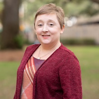 A smiling woman with short hair wearing a maroon cardigan and a patterned shirt, standing outdoors in a park setting.