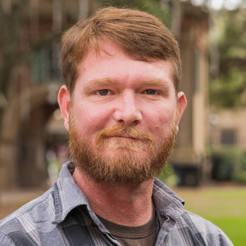 Headshot of James Lohmar, slightly smiling man with red hair and beard against an outdoor campus background.
