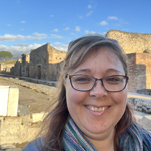 Head shot of Allison Sterrett Krause, a smiling woman with glasses in fron of the preserved building of Pompeii.