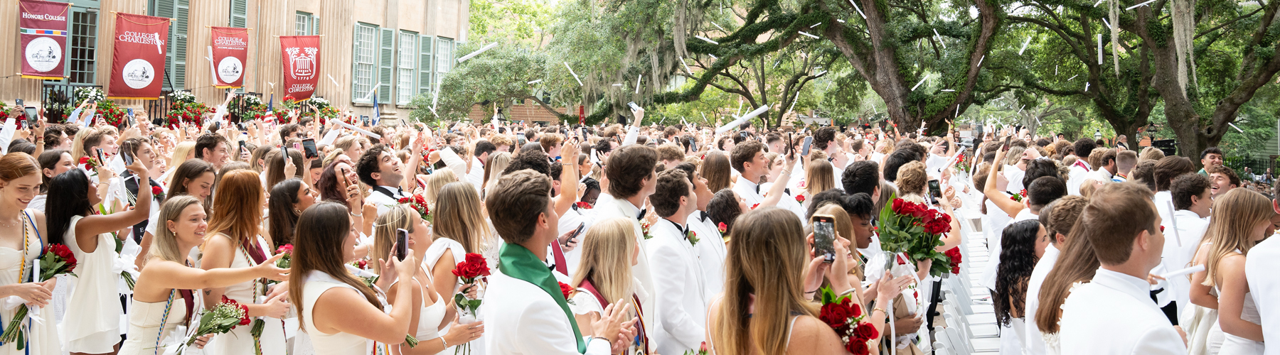 Large crowd of graduates in white holding red rose bouquets and cheering beneath large oak trees at an outdoor commencement.