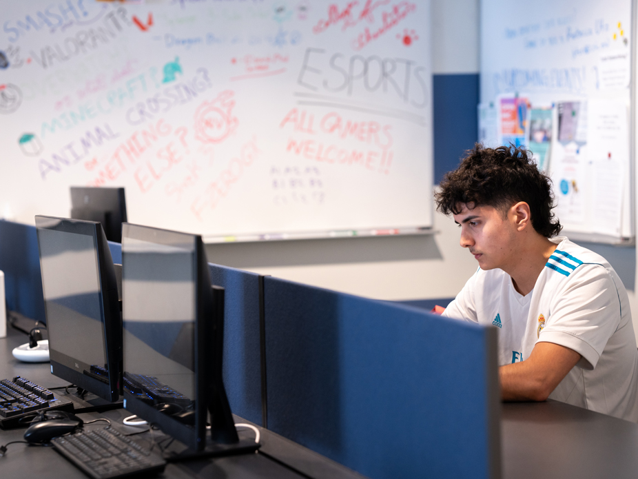 A student works at a computer station with an ESports organizational message on the whiteboard in the background.