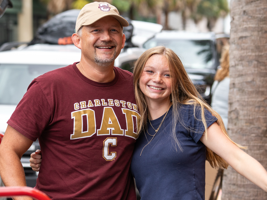A dad wearing a "Charleston Dad" t-shirt and College of Charleston hat smiles at the camera with his College of Charleston daughter.
