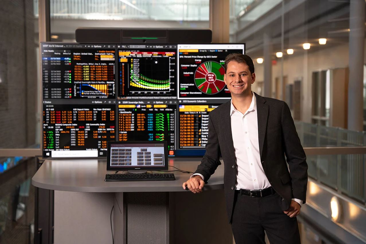 student standing in front of computer screens showing finance graphs