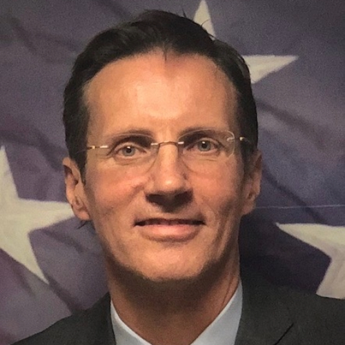 A man with glasses smiling in front of a flag