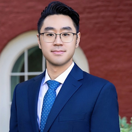 A man with glasses and dark hair smiling wearing a business suit