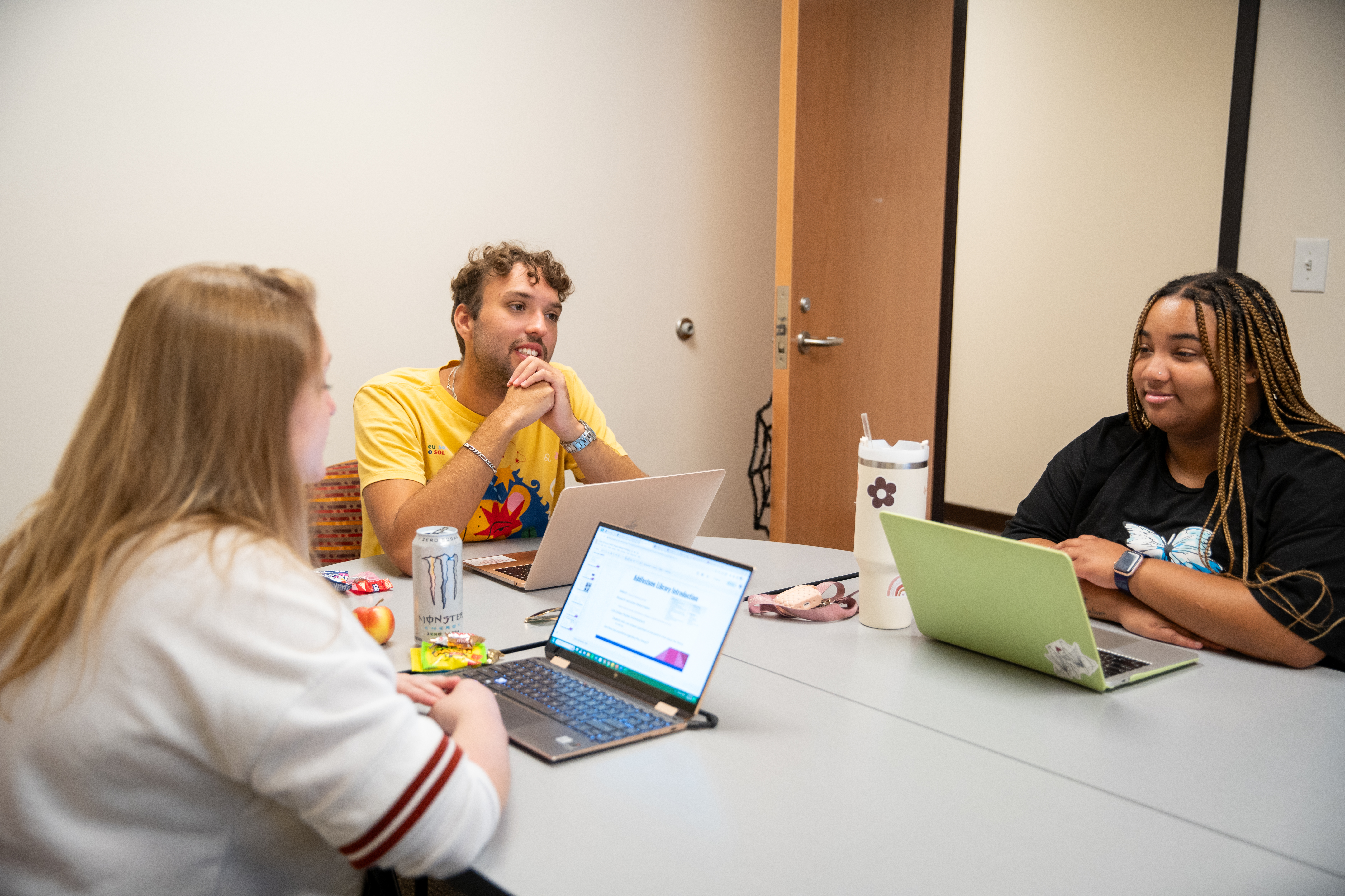 Three students collaborate around a table with laptops in a small meeting room.