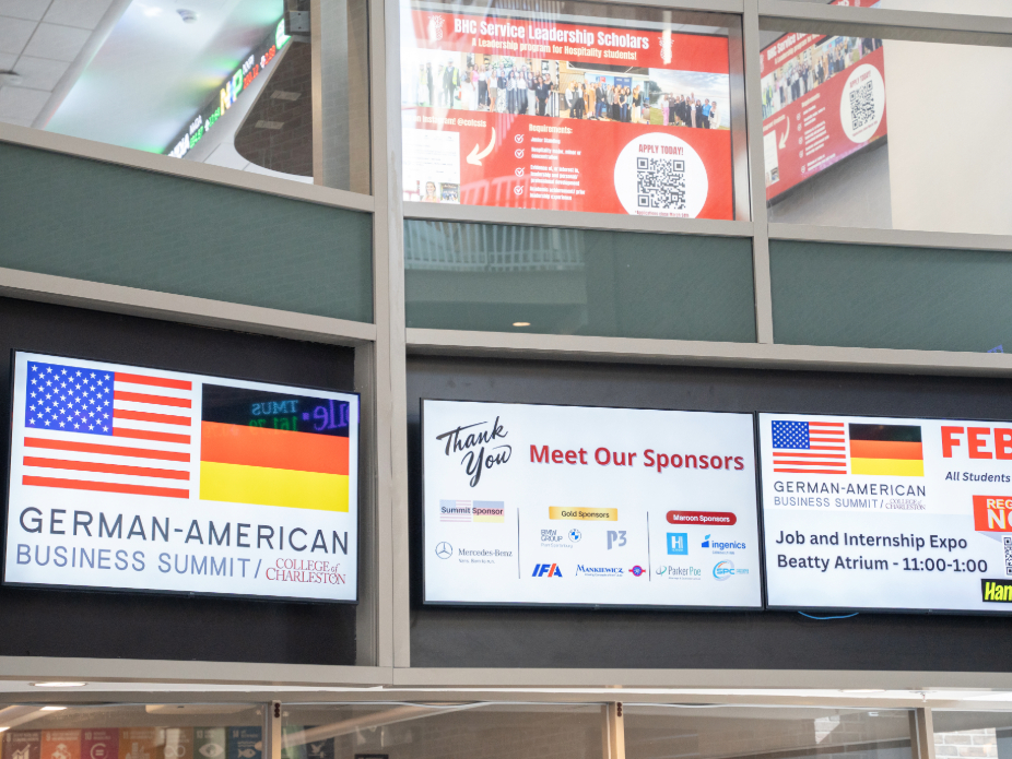 Photo of tv screens in venue space of the expo that showcases German-American Business Summit graphic and sponsor logos.