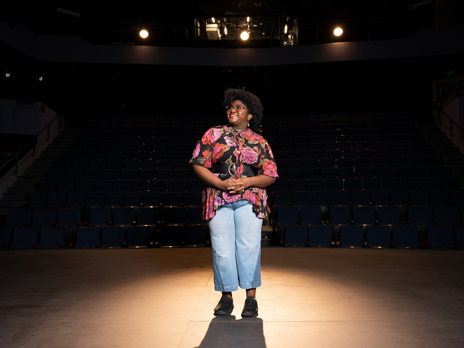 A person stands on a theater stage under bright overhead lights, facing forward with hands clasped. They are wearing a colorful floral shirt, light blue cropped jeans, and black shoes. Rows of empty dark blue seats fill the background, and the stage lighting creates a dramatic spotlight effect.
