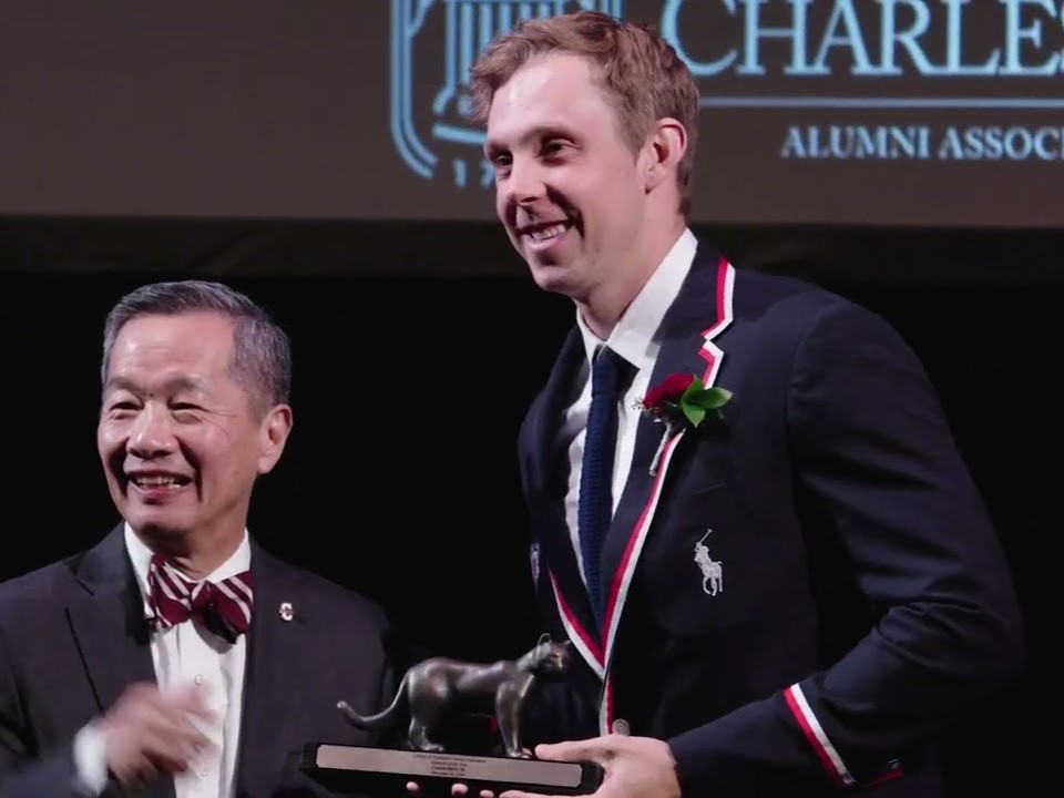 A young man wearing a Team USA Olympic blazer stands holding a small award alongside a man in a black suit and bowtie. 