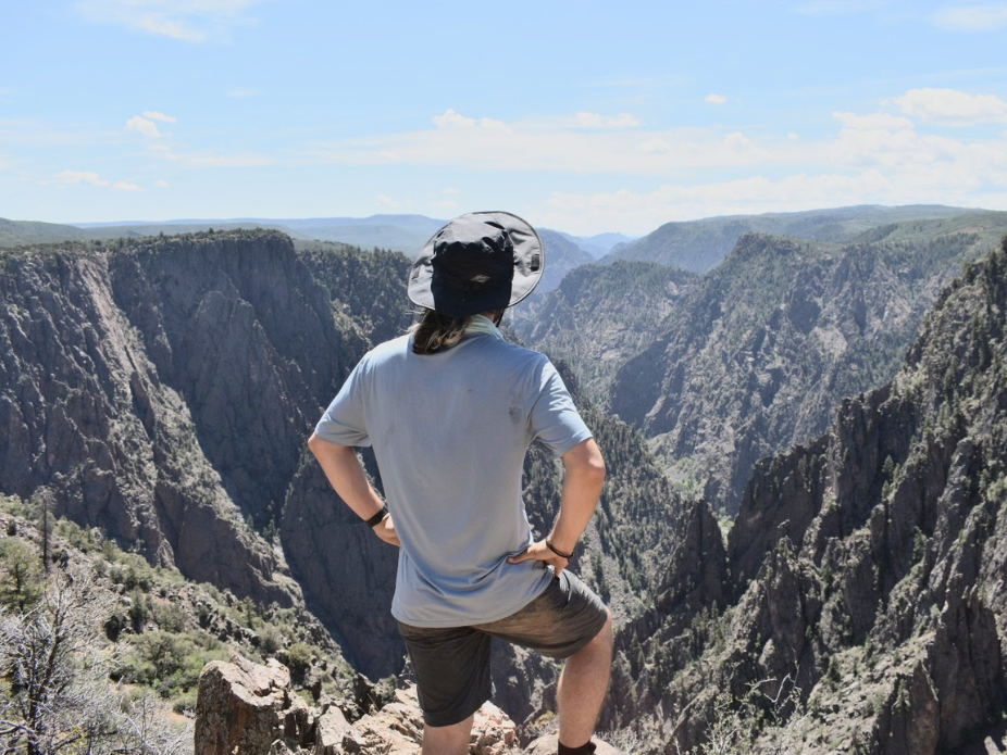 Person wearing a wide‑brim hat stands on a rocky overlook, looking out over a deep canyon with steep cliffs and distant mountains under a blue sky.