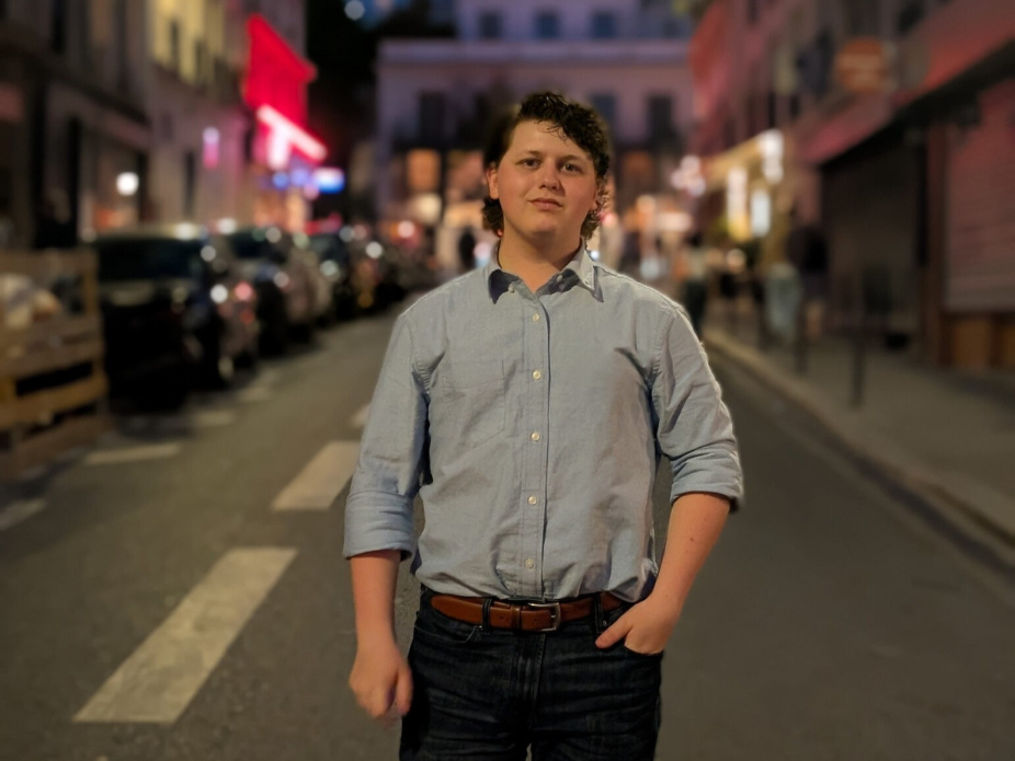 A person stands in the middle of a city street at night, wearing a light blue button‑down shirt and dark jeans, with softly blurred buildings and streetlights in the background.