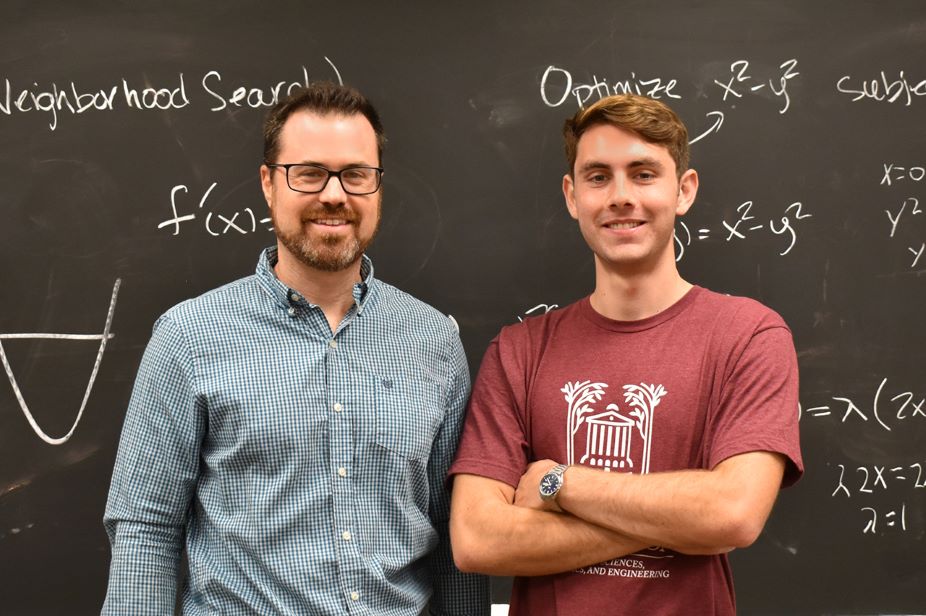 Assistant professor Dan Maroncelli poses beside statistics major Aidan Riordan in front of a chalkboard.