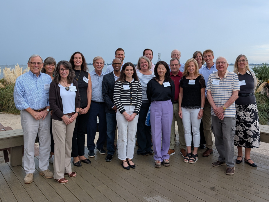 A group of 18 adults poses in front of a seascape.