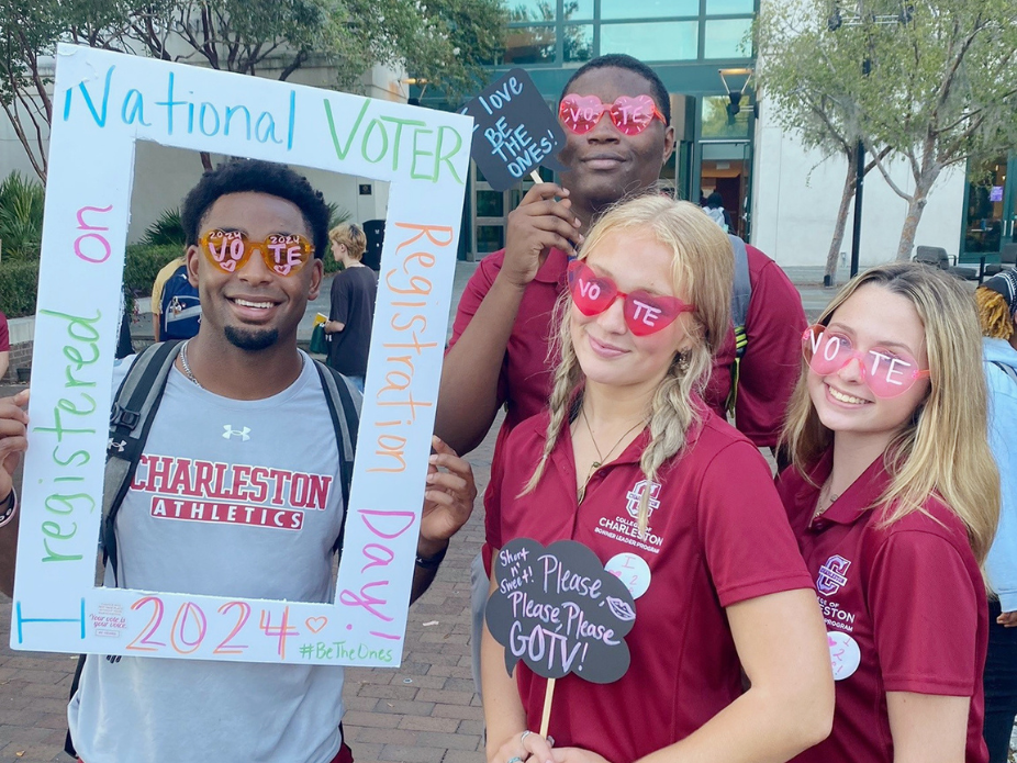 Four college students pose for a photo, each holding signs that encourage their peers to register to vote.