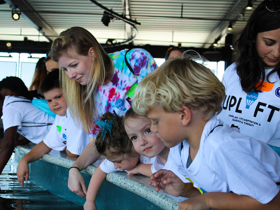 A young female volunteer assists a collection of young children who are peering over the edge of a large, open-air aquariuam tank.